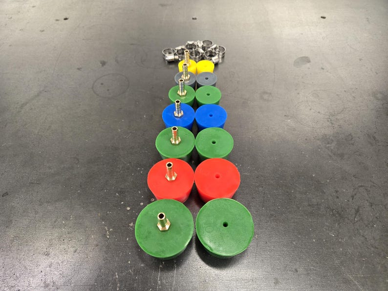 May include: An assortment of colorful rubber grommets with brass fittings, arranged in a vertical line. The grommets are in various colors, including green, red, blue, yellow, and gray. A collection of metal clamps is at the top.