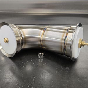 May include: A stainless steel pipe with a curved design, featuring white end caps and brass fittings. A small metal clamp sits nearby. The pipe has visible weld marks and is designed for industrial use.