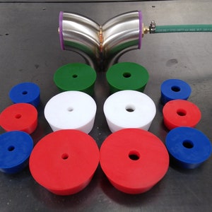 May include: A set of 12 rubber plugs in various sizes and colors, including red, white, blue, and green. The plugs have a central hole and are arranged in a semi-circle pattern on a gray surface. A stainless steel pipe fitting is also visible in the image.
