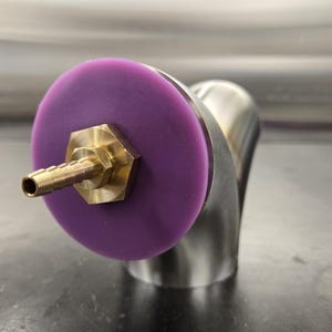 May include: A close-up of a metal component with a purple disc and a brass fitting. The component has a curved, polished metal body. The brass fitting has a hose barb. The purple disc is circular.