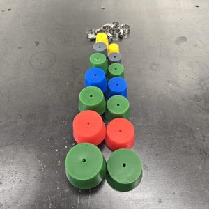 May include: An assortment of colorful, cylindrical objects in red, green, blue, yellow, and gray, with a small hole in the center of each. Several silver metal clamps are also present. The objects are arranged in a vertical line.
