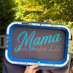 May include: A rectangular blue embroidery hoop holding a piece of dark gray fabric. The fabric features the word Mama embroidered in bright teal thread with a pink outline, and the text Shore Comfort LLC embroidered in a smaller white font.
