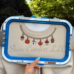 May include: A beige sweatshirt secured in a blue 8x13 inch embroidery hoop, featuring an embroidered gold chain necklace design with charms including an acorn, pumpkin, coffee mug with a heart, maple leaf, mushroom, and pinecone.
