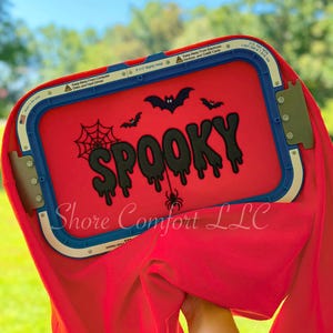 Puede incluir: Una tela roja sujeta en un bastidor de bordado rectangular azul y blanco. La tela presenta el texto bordado en negro que dice SPOOKY, con un diseño de telaraña, tres murciélagos y una araña colgante alrededor de las letras.