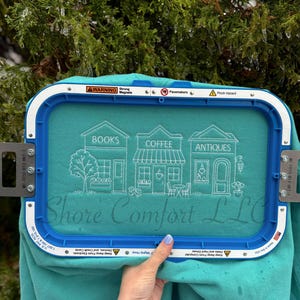 May include: Teal sweatshirt featuring an embroidered design of a bookstore, coffee shop, and antique shop. The design is framed by a blue embroidery hoop. The text "Shore Comfort LLC" is also visible.
