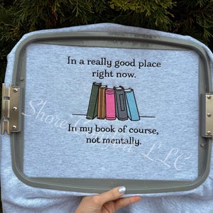 以下が含まれることがあります： 「In a really good place right now. In my book of course, not mentally.」のテキストが入ったライトグレーのスウェットシャツ。カラフルな本が並んだデザイン。スウェットシャツはグレーの刺繍枠に収められています。