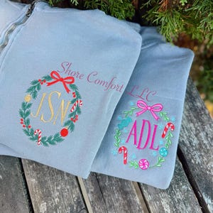 May include: Two light blue zip-up sweatshirts with embroidered holiday wreath designs. One wreath features the initials "JSN" in gold, and the other has "ADL" in pink. The text "Shore Comfort LLC" is also visible.