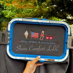 Könnte beinhalten: Ein dunkelgrauer Stoff in einem blauen Stickrahmen mit gestickten Symbolen einer Eistüte, einer amerikanischen Flagge, einer Sodaflasche und herzförmigen Sonnenbrillen. Darunter steht der Text stars, stripes & summertime und Shore Comfort LLC.