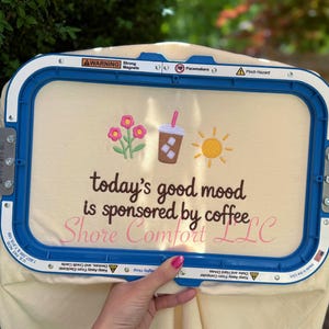 Puede incluir: Tela color crema con bordados de flores, una bebida de café y un sol. El texto dice "today's good mood is sponsored by coffee" y "Shore Comfort LLC". La tela está sujeta en un bastidor de bordado azul.