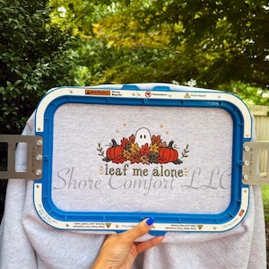 May include: A light gray sweatshirt secured in a blue rectangular embroidery hoop. The fabric features an embroidered design of a white ghost surrounded by orange pumpkins and autumn leaves, with the text "leaf me alone" stitched below.
