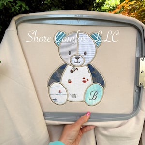 Può includere: Felpa beige ricamata con un design a orsetto patchwork. L'orso ha accenti blu, grigi e bianchi, con una iniziale turchese "B" sul piede. È visibile anche il testo "Shore Comfort LLC".