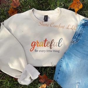 May include: Cream-colored sweatshirt with the embroidered text "grateful for every little thing" in fall colors. The brand name "Shore Comfort LLC" is embroidered above the main text. A pair of blue jeans is visible on the right.