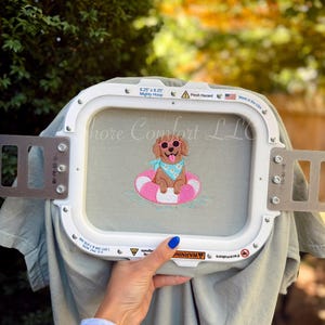 May include: A white rectangular embroidery hoop holding sage green fabric with an embroidered design of a brown dog wearing pink sunglasses and a light blue bandana, sitting in a pink and white striped inner tube floating on blue water.