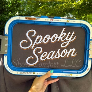 May include: A blue rectangular embroidery hoop holding dark gray fabric with the white and orange embroidered text Spooky Season. The frame features warning labels regarding strong magnets and pacemakers, along with the text Shore Comfort LLC.