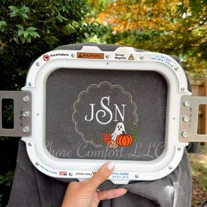 May include: A dark gray fabric held in a white rectangular embroidery hoop. The fabric features an embroidered design with the letters JSN inside a scalloped circle, accompanied by a small white ghost, an orange pumpkin, and autumn leaves.