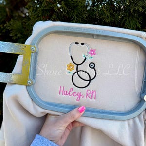 May include: A beige sweatshirt with an embroidered stethoscope design, pink flowers, and the name "Haley, RN" in pink thread. The sweatshirt is held in an embroidery hoop.