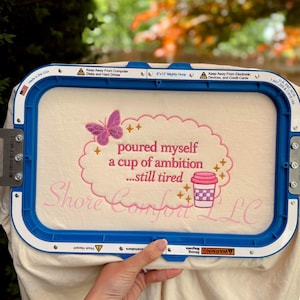 May include: A cream-coloured fabric is held in a blue embroidery hoop. The fabric features pink embroidered text and a butterfly design. Text reads "poured myself a cup of ambition...still tired" and "Shore Comfort LLC".