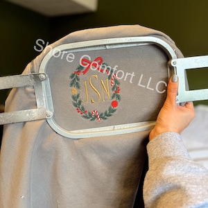 May include: A gray sweatshirt with an embroidered Christmas wreath design. The wreath includes red bows, candy canes, and gold initials. The text "Shore Comfort LLC" is visible.