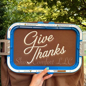 May include: A brown fabric piece secured in a blue rectangular embroidery hoop. The fabric features the embroidered text "Give Thanks" in a cream-coloured script font, with the smaller text "ShoreComfort LLC" printed below in a light grey.