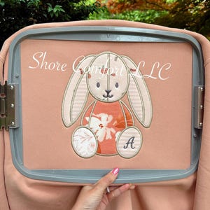 May include: A peach-coloured sweatshirt featuring an embroidered bunny design. The bunny has long ears, a floral patterned dress, and the letter "A" on its foot. The text "Shore Comfort LLC" is above the bunny.