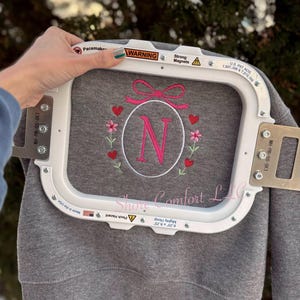May include: Gray sweatshirt with a pink embroidered initial "N" inside a floral wreath with hearts and a bow. The sweatshirt is held in a white embroidery hoop. The text "Shore Comfort LLC" is visible.