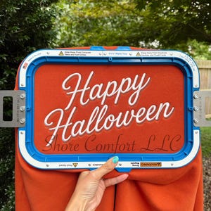 May include: A rectangular blue embroidery hoop securing a piece of orange fabric. The fabric features white embroidered text that reads Happy Halloween, with the text Shore Comfort LLC printed in a dark script font below the embroidery.