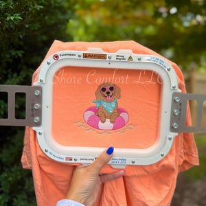 May include: A peach-coloured t-shirt featuring an embroidered design of a brown dog wearing pink sunglasses and a light blue bandana with white flowers, sitting in a pink and white striped inner tube floatie on a white embroidery hoop frame.