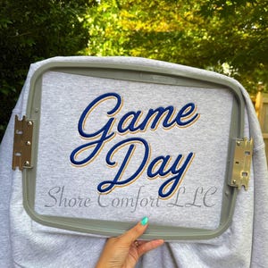 May include: A light grey sweatshirt secured in a plastic embroidery hoop. The fabric features the embroidered text "Game Day" in blue with a gold outline. Below the main text, the words "Shore Comfort LLC" are embroidered in a grey script font.