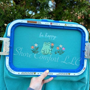 May include: A teal fabric is held in a blue embroidery hoop. The fabric features embroidered text that says "be hoppy" and "Shore Comfort LLC". A green frog and colourful flowers are also embroidered on the fabric.