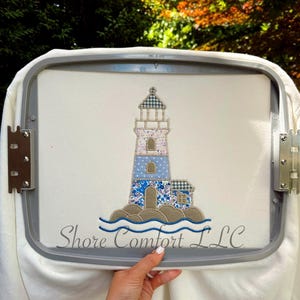 May include: An embroidery design of a lighthouse on white fabric secured in a gray hoop. The lighthouse features blue, white, and tan patterned sections, sitting on tan rocks with blue waves. The text Shore Comfort LLC is embroidered below.