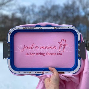 Puede incluir: Una sudadera rosa se sostiene en un bastidor de bordado. La sudadera tiene el texto bordado "just a mama in her string cheese era" en hilo rosa. El bastidor es azul y blanco.