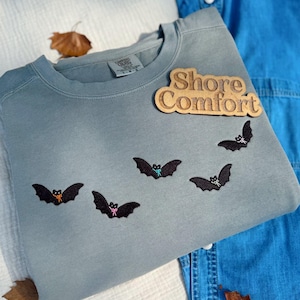May include: A light blue sweatshirt with embroidered black bats, each adorned with a colorful bow. The wooden text "Shore Comfort" is attached to the sweatshirt. A denim shirt is visible in the background.