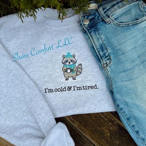 May include: A pale grey sweatshirt with the text "Shore Comfort LLC" and "I'm cold & I'm tired." An embroidered raccoon wearing a hat and scarf is holding a mug. Light blue denim jeans are next to the sweatshirt.