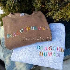 Puede incluir: Se muestran dos sudaderas, una marrón y otra gris claro. Ambas tienen la frase bordada "BE A GOOD HUMAN" en letras coloridas. La sudadera marrón está encima, con la gris claro debajo. También es visible el nombre de la marca "Shore Comfort LLC".