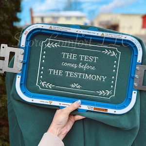 May include: A teal green sweatshirt held in an embroidery hoop. The text "THE TEST comes before THE TESTIMONY" is embroidered in white. The hoop is blue with metal components. The background is blurred, showing a building and trees.