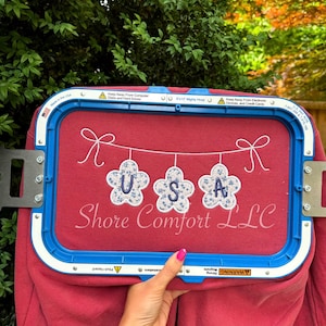 Könnte beinhalten: Ein rotes Sweatshirt mit einem blau-weißen Blumendesign mit der Aufschrift "USA" wird in einem blauen Stickrahmen gehalten. Der Text "Shore Comfort LLC" befindet sich darunter. Der Rahmen ist mit "Made in the USA" beschriftet.