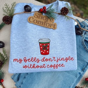 May include: A light blue sweatshirt with a red and green coffee cup design and the phrase "my bells don't jingle without coffee." A wooden tag with "Shore Comfort" is attached. The sweatshirt is folded on top of blue jeans.
