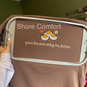 May include: Brown sweatshirt with the text "Shore Comfort" and "you know why I'm here". The design includes an embroidered image of deviled eggs with flowers. The sweatshirt is held in a metal embroidery hoop.