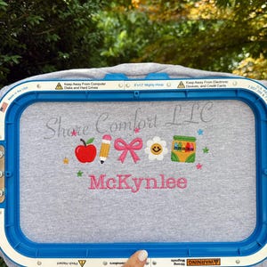 Peut inclure: Un vêtement en tissu gris fixé dans un cadre à broder bleu de 20,32 x 33,02 cm. La broderie présente le texte Shore Comfort LLC, le nom McKynlee en rose, et des icônes colorées dont une pomme rouge, un crayon, un nœud rose, une fleur et des crayons.