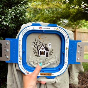 May include: A light olive green shirt featuring an embroidered design of a small house, a ghost, a crescent moon, stars, and a bare tree. The fabric is secured within a blue plastic Mighty Hoop embroidery frame with visible warning labels.