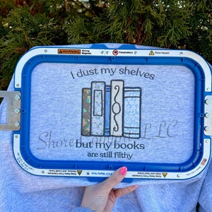 May include: A light gray sweatshirt with the text "I dust my shelves but my books are still filthy" and a design of four embroidered books in various patterns. The sweatshirt is held in a blue embroidery hoop.