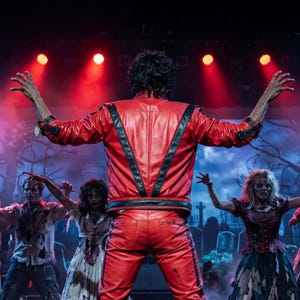 May include: A bright red leather jacket with black V-shaped accents on the back and sleeves, worn by a person with dark curly hair standing with arms outstretched on a stage, surrounded by performers in tattered clothing under red spotlights.