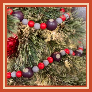 May include: A Christmas tree decorated with a garland of wooden beads in shades of red, gray, and dark purple. The garland is draped across the green pine needles, with a red, faceted ornament visible.