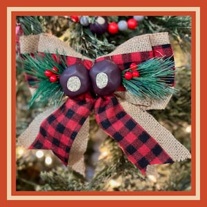 May include: A festive Christmas tree bow with a rustic design. The bow features a burlap base, accented with red and black buffalo plaid ribbon. Two dark brown, round ornaments with gold centers are at the bow's center, with faux greenery and red berries.