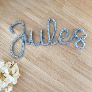 Peut inclure: Le mot "Jules" en bleu clair, tricoté en écriture cursive. Les lettres sont faites d'un fil épais et texturé et sont affichées sur une surface en bois clair. Un arrangement de fleurs séchées est dans le coin inférieur gauche.