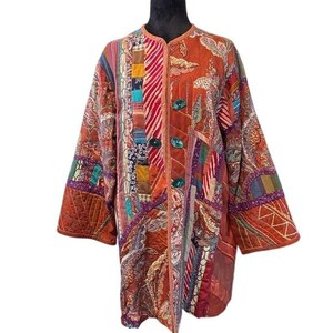 May include: A patchwork coat in shades of orange, red, and purple. The coat features a variety of patterned fabrics, including paisley and floral designs. It has a round collar, long sleeves, and large green buttons. The coat is quilted and has a relaxed fit.