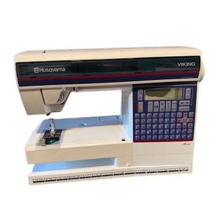 May include: A white Husqvarna Viking sewing machine with a blue and red accent stripe. The machine has a digital display and a keyboard of buttons. The model name is Rose.