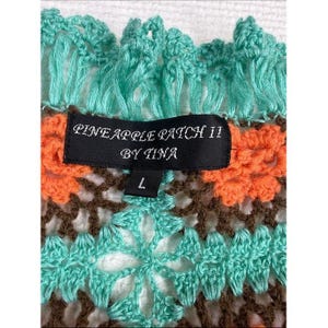 May include: A crochet garment featuring a vibrant pattern of turquoise, orange, and brown yarn. A black rectangular label is stitched to the center, displaying the white text PINEAPPLE PATCH II BY TINA and the size L on a smaller tab below.
