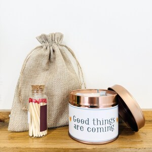 May include: A rose gold metal candle tin with a white label reading Good things are coming, a small glass bottle filled with matches featuring red tips and a cork stopper, and a beige burlap drawstring pouch with a woven texture and rope ties.