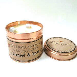 May include: A copper candle tin with a brown label that reads "Congratulations on your engagement Daniel & Rose". The candle is white and has three white wax cubes on top. The lid of the tin is copper and has a brown label that reads "Fresh Linen" with a heart symbol. The label also says "Place candle on the lid when burning."